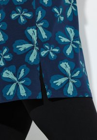 Marineblå tunika med stora teal-blommönster, korta sidofickor och en mjuk textur, tillsammans med svarta leggings.