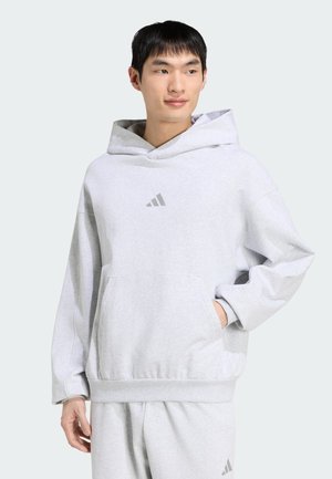 Młody mężczyzna noszący jasnoszary kaptur Adidas i pasujące spodnie dresowe, stoi z jedną ręką w kieszeni, patrząc na bok.