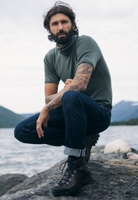 Grå kortærmet skjorte, mørk denim bukser og brune vandrestøvler med sorte snørebånd. Mand, der knæler på sten ved vandet, viser arm tatoveringer.