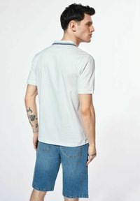 Biała koszulka polo męska z krótkimi rękawami, z kontrastującym granatowym kołnierzykiem, zestawiona z niebieskimi szortami jeansowymi. Widoczne tatuaże na ramieniu.