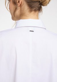 Valkoinen nappikauluspaita, jossa on klassinen kaulus ja pieni metallinen brändilogo takana. Sujuva pinta ja minimalistic design.