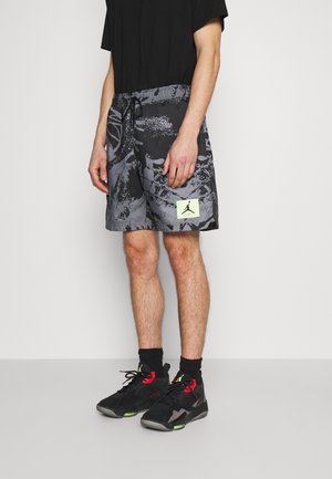 Mężczyzna noszący czarne i szare krótkie spodenki z wzorem oraz naszywką z logo Jordan, czarną koszulkę, czarne skarpetki i czarno-czerwone sneakersy.