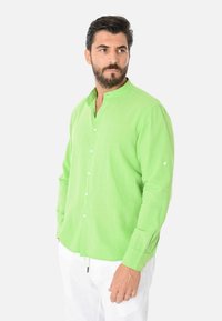 Camicia a maniche lunghe verde in lino con colletto a band e chiusura frontale a bottoni, dotata di bottoni ai polsini. Abbinata a pantaloni bianchi, per un look rilassato.