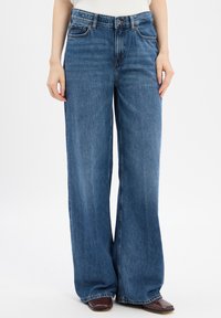 Brede spijkerbroeken van denim in een middenblauwe was, met vijf zakken, riemlussen en subtiele slijtageplekken. Materiaal is zachte katoen met een gladde textuur.