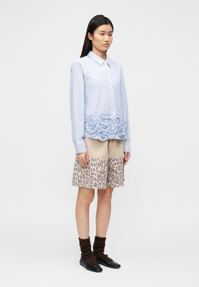 Ανοιχτό μπλε πουκάμισο με κουμπιά και βολάν στο τελείωμα, συνδυασμένο με μπεζ σορτς με σχέδιο λεοπάρ. Μαύρα παπούτσια και καφέ κάλτσες.