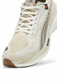 Beige idrottssko med övre del av mesh, vita snören och gummisula. Har ett texturerat mönster och en logotyp på sidan.