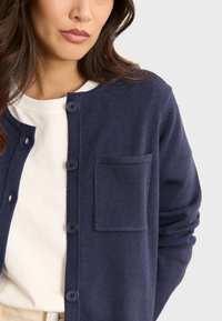 Marinblå kofta i mjukt stickat material. Har rund hals, knäppning med knappar och en enda bröstficka. Slät textur, avslappnad passform.