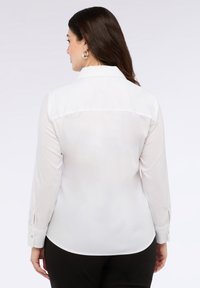 Biała bluzka z długim rękawem o gładkiej fakturze i mankietach zapinanych na guziki, z karczkiem w tylnej części. Noszona z czarnymi spodniami.