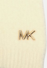 Kremowy dzianinowy materiał o miękkiej fakturze, z wyraźnym złotym logo "MK". Wzór jest prosty i elegancki.