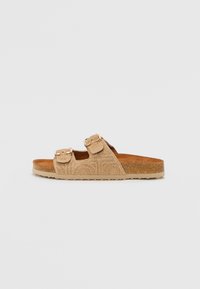 Sandale slide din suede bej, cu două curele reglabile cu catarame aurii, un suport din plută și un material texturat. Talpă plată cu un ușor profil.