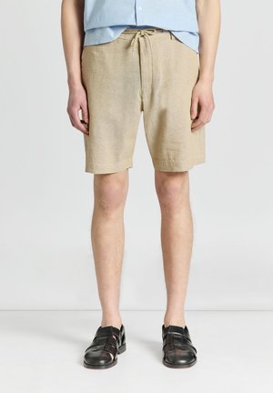 Uomo con pantaloncini beige con coulisse, camicia azzurra e sandali neri in pelle stile pescatore, in piedi su pavimento bianco.