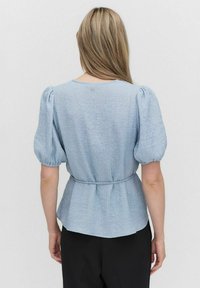 Ljusblå blus med puffärmar och texturerad yta, midjeinsnörning och knapprad bak. Matchas med svarta byxor.