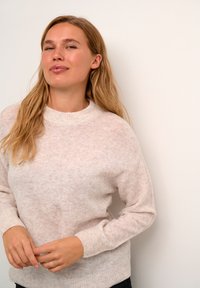 Svetlo roza, prevelik pulover z teksturiranim pletenjem, visokim ovratnikom in rebrastimi manšetami, v kombinaciji z črnimi hlačami. Prisotni so subtilni barvni odtenki.