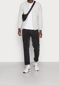 Veste bomber grise texturée portée par-dessus un t-shirt blanc, jean noir et baskets blanches. Un sac bandoulière noir est porté.