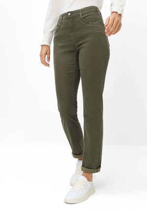 Femme portant un pantalon vert olive retroussé, des baskets blanches et un haut blanc à manches longues, debout devant un fond blanc.