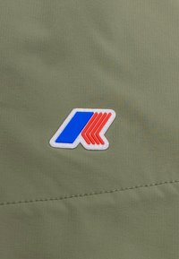 Punto logo su tessuto verde oliva con un design blu e rosso caratterizzato da forme angolari e un bordo bianco, cucito sul materiale.