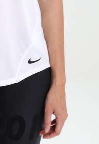 Witte sporttop met korte mouwen, met een zwart Nike-logo aan de linkerkant; gecombineerd met zwarte leggings met grote tekst.