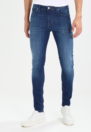 Homme portant un jean slim bleu et des baskets blanches, debout devant un fond blanc uni.