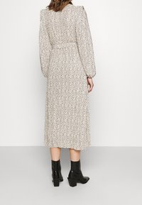 Krémové šaty s černým puntíkovaným vzorem, dlouhými rukávy a staženým pasem. Modelka má na sobě černé kotníkové boty na podpatku.