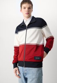 Giacca con zip che presenta blocchi di colore blu navy, rosso e bianco, con polsini e colletto a coste. Mostra una toppa con logo sulla parte anteriore inferiore.
