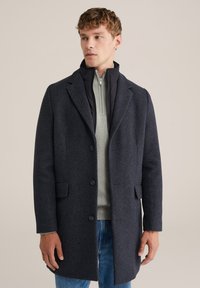 Cappotto in misto lana blu navy con un design strutturato, a un petto, e due bottoni. Indossato sopra un maglione grigio chiaro con zip.