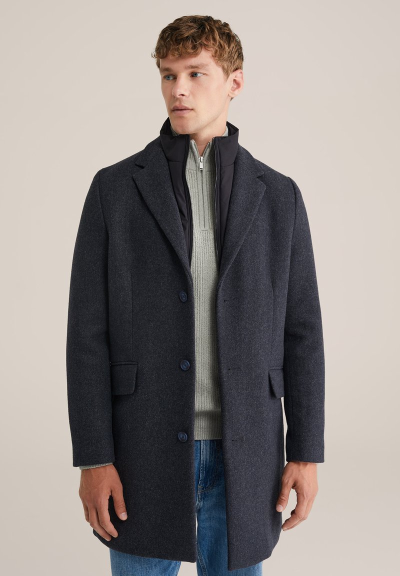 Cappotto in misto lana blu navy con un design strutturato, a un petto, e due bottoni. Indossato sopra un maglione grigio chiaro con zip.