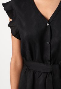 Robe noire avec un tissu texturé, col en V, manches courtes à volants, et une ceinture en tissu. Fermée par des boutons noirs à l'avant.