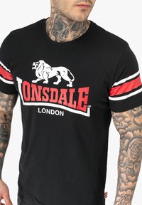 T-shirt nera con logo di un leone bianco e testo "LONSDALE LONDON" in rosso. Maniche corte con strisce rosse e bianche. Materiale in cotone.
