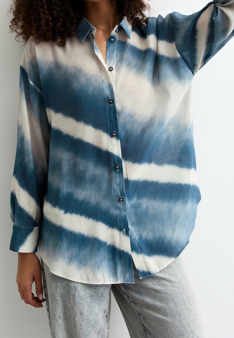 Chemise ample à manches longues boutonnée avec motif tie-dye bleu et blanc horizontal portée avec un pantalon gris clair.
