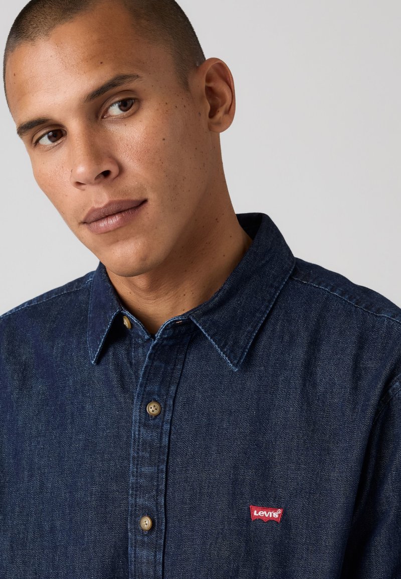 Jeune homme aux cheveux courts portant une chemise en jean Levi's bleu foncé, regardant légèrement sur le côté sur un fond uni.