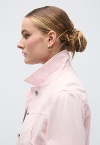 Ροζ τζιν μπουφάν με ψηλό γιακά και ασημένια κουμπιά, συνδυασμένο με χρυσούς κρίκους σκουλαρίκια, απεικονίζεται πλαγίως σε ουδέτερο φόντο.