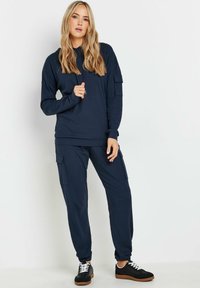 Granatowy dres z prążkowanej tkaniny, składający się z bluzy na zamek z długimi rękawami i kieszeniami bojowymi, w zestawie z dopasowanymi spodniami i czarnymi trampkami.