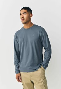 Long-sleeve, crewneck shirt in muted blue fabric. Features a subtle logo on the chest. Paired with light beige pants, showcasing a relaxed fit.