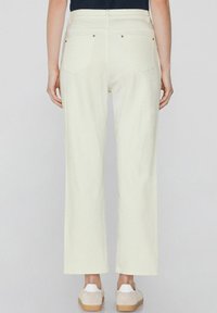 Pantalones de pierna ancha, de color crema claro, hechos de una tela suave. Presentan bolsillos traseros y acentos de metal. Combinados con zapatos beige tipo slip-on.
