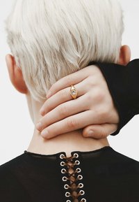 Anillo de oro con una piedra facetada, llevado en una mano que descansa sobre un cuello. La persona tiene el cabello corto y de color claro, y lleva una blusa negra con cordones.