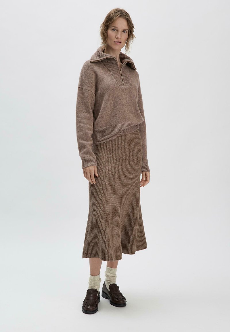 Brauner Strickpullover mit hohem Kragen und Reißverschluss, kombiniert mit einem passenden gerippten Midi-Rock, getragen mit cremefarbenen Socken und dunklen Schuhen.