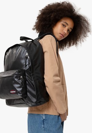 Black Eastpak backpack with a shiny finish, two front zip pockets, and padded shoulder straps, worn over a beige jacket against a white background.