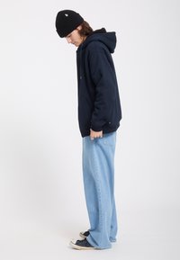 Jeune personne portant un bonnet noir, un sweat à capuche bleu marine, un jean large bleu clair et des baskets noires, debout de côté avec la tête légèrement baissée.