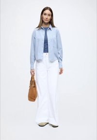 Cămașă albastru deschis cu nasturi peste un top albastru, asortată cu pantaloni largi albi. Ține o geantă maro și stă într-un cadru neutru.