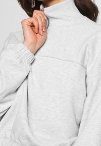 Sudadera de cuello con cremallera en gris claro, con una textura suave, puños elásticos y detalles de costura en el pecho. Mano que tira de la cremallera ligeramente.