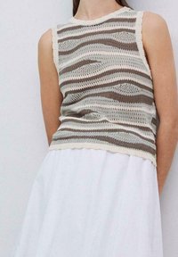 Gebreide zonder mouwen top in crème, beige en taupe. Bevat een golfpatroon met een geschulpte rand. Gaat over een vloeiende witte jurk.