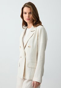 Cremfarbener Leinenblazer mit doppelter Knopfleiste, abgerundeten Revers und zwei vorderen Taschen. Mit hellfarbenen Knöpfen und einem lässigen Schnitt.