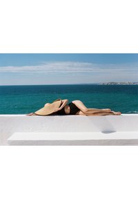 Black swimsuit, large straw sunhat, bronze skin. Lying on a white ledge with ocean view; horizon is clear blue with few clouds.