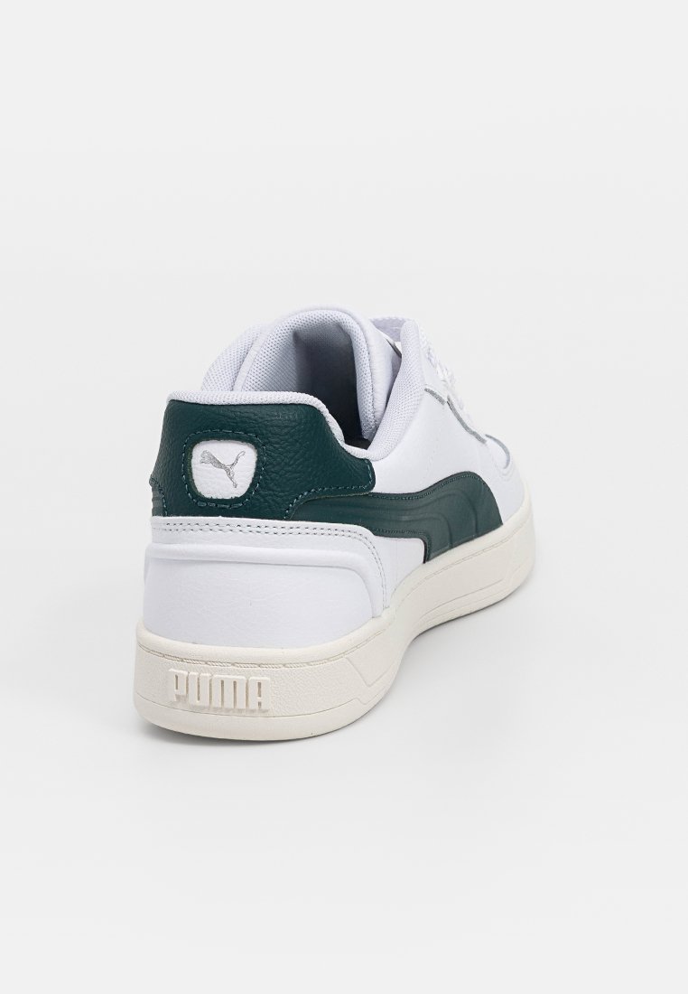 Scarpa sportiva bianca con un accento in pelle verde, tacco testurizzato e lacci bianchi. Presenta un logo rotondo e la scritta "PUMA" impressa sulla suola.
