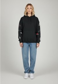 Felpa nera con cappuccio e tasca frontale, caratterizzata da grafiche e motivi multicolori sulle maniche. Abbinata a jeans azzurri chiari e scarpe bianche.