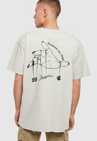 Valge puuvillane T-särk, millel on must graafika käest, mis hoiab nööre, ning tekst "99" ja "Rocawear". Lahti istuv, lühikeste varrukatega, ümar kaelus.