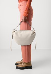 Person wearing pink pants and tan boots holds large off-white handbag with silver chain handle against white background.