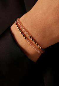 Roze kraaltjesarmband met gouden accentkralen, gelaagd op een pols tegen een donkere achtergrond, waardoor textuur en subtiele glans worden getoond.