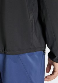 Μαύρο μπουφάν με λείο, υφασμάτινο υλικό, που διαθέτει ρυθμιζόμενο στρίφωμα και μπλε παντελόνι από κάτω. Λεπτομέρεια περιλαμβάνει κορδόνι ρύθμισης στο κάτω μέρος.