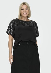 Czarna bluzka z cekinowym akcentem na ramieniu i krótkimi rękawami, w połączeniu z czarną spódnicą o wysokim stanie. Gładka struktura materiału.
